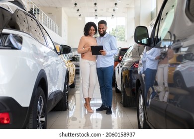 Young Black Couple Choosing Car In Modern Dealership Center, Holding Clipboard And Checking Vehicle Characteristics In Catalog, Happy African American Spouses Buying Automobile In Showroom