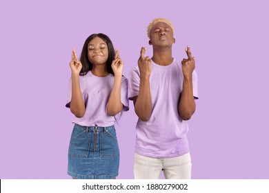 Young Black Couple Crossing Fingers And Making Wish With Closed Eyes, Begging For Dream To Come True On Lilac Studio Background. Hopeful African American Woman With Her Boyfriend Praying For Good Luck