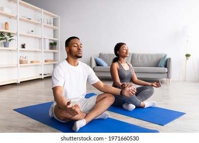 Young black couple meditating with closed eyes, sitting on sports mats in lotus pose at home, full length. African American man and his girlfriend doing daily yoga routine, practicing mindfulness