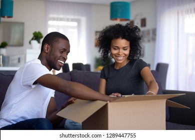 Young Black Couple Open Cardboard Box Or Parcel Unpacking Sitting On Couch In Living Room On Moving Day, Happy African Man And Woman Packing Belongings, Relocation Removals Settle In New Home Concept