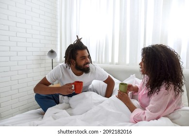 Young Black Couple Talking And Drinking Tea Or Coffee In Bed At Morning. Concept Of Relationship And Spend Time Together. Idea Of Domestic Lifestyle. Man And Woman At Spacious Bedroom In Modern Flat