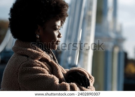 Young black curly woman looking away with determination. She has a blurry metallic bridge on the background. Winter cold morning.