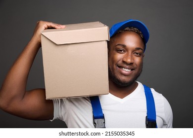 A Young Black Delivery Man In A Blue Baseball Cap Holding And Carrying A Cardboard Box On His Shoulder. With One Hand, Strong Dark-skinned Courier In Work Overalls On Gray Background Holds A Parcel