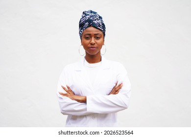 Young black doctor or scientist stands outside against a white background wearing a lab coat and a head wrap turban with her arms crossed and smiling                               