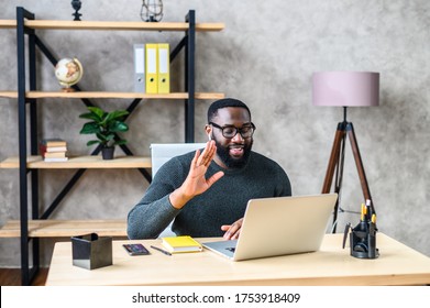 Young Black Employee Is Using Video Connection For Virtual Meeting With Co-workers, A Guy Sits At The Desk In Contemporary Office, Greeting And Waving Into Webcam