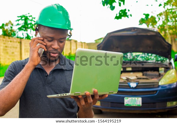 Young black engineer making a phone call and also\
holding his laptop checking out something with a broken down car\
behind him