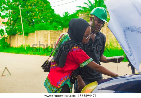 Young\
black engineer using his laptop to examine a car and also showing\
an African woman something in the engine of the\
car