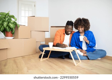 Young Black Family Couple Planning Shopping For New Home, Making Checklist While Sitting Among Cardboard Boxes In Their Flat, African American Spouses Drinking Coffee And Writing In Notepad - Powered by Shutterstock