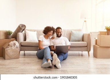 Young Black Family Couple Using Laptop Sitting On Floor Indoor After Moving New Home. Real Estate, House Ownership And Relocation Concept. Spouses Browsing Internet Sitting Among Cardboard Boxes