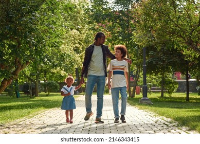 Young Black Family Walking On Pavement Road In Green Park. Father Hug Son And Hold Hand Of Little Daughter. Family Relationship And Spending Time Together. Fatherhood And Parenting. Sunny Summer Day