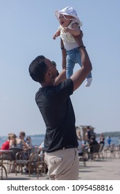 Young Black Father With Aviator Sunglasses And Black Shirt Lifting Cute Little Baby Girl With Hat Up In The Air On A Summer Day