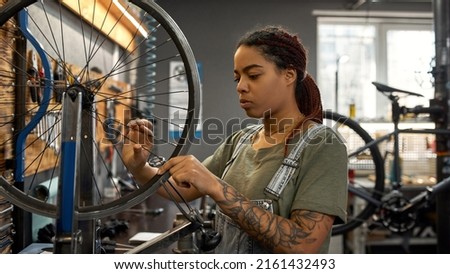 Young black female cycling technician checking bicycle wheel spoke with bike spoke key in modern workshop. Bike service, repair and upgrade. Garage interior with tools and equipment