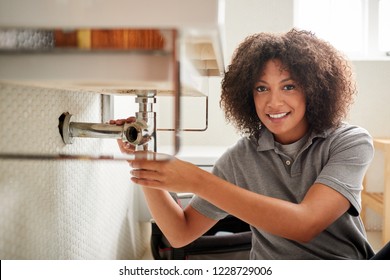 Young Black Female Plumber Sitting On The Floor Fixing A Bathroom Sink, Looking To Camera