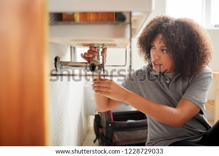 Young black female plumber sitting on the floor fixing a bathroom sink, seen from doorway