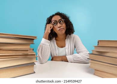 Young black female student in glasses sitting at desk with piles of books, daydreaming, feeling bored during lesson on blue studio background. African American woman preparing for dull college exam - Shutterstock ID 2149539427