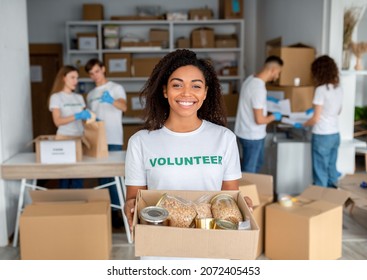 Young black female volunteer holding food donation box and smiling to camera while posing to camera in charity office working at volunteering organization - Powered by Shutterstock