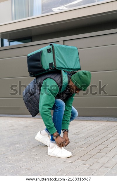 A young black food delivery man in a green uniform\
walks up to the house.