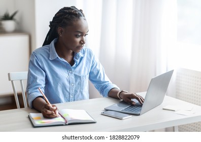 Young Black Freelancer Woman Working On Laptop At Home Office And Taking Notes, Sitting At Desk Near Window, Looking At Screen, Writing Down Information From Computer, Enjoying Remote Work, Copy Space