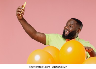 Young black gay man 20s in green t-shirt bow tie hold bunch of air inflated helium balloons celebrating birthday party doing selfie shot on mobile cell phone isolated on plain pastel pink background.