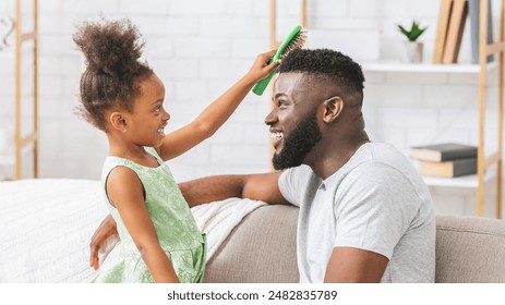 A young black girl with curly hair is brushing her fathers hair with a green brush while he sits on a couch. The girl is smiling and looking at her father, while he is smiling and looking at her. - Powered by Shutterstock