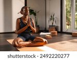 Young black girl practicing yoga indoors, sitting cross-legged on a yoga mat with her hands in a prayer position. She appears calm and focused, with plants and yoga blocks in the background.