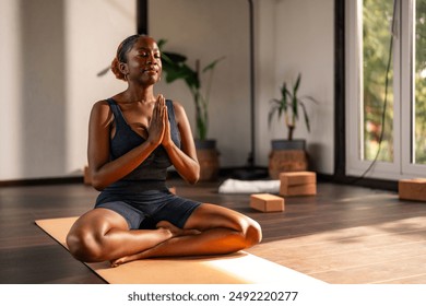 Young black girl practicing yoga indoors, sitting cross-legged on a yoga mat with her hands in a prayer position. She appears calm and focused, with plants and yoga blocks in the background. - Powered by Shutterstock