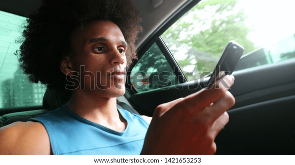Young black guy in backseat of taxi riding car\
while holding smartphone