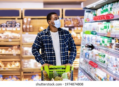 Young black guy in disposable face mask selecting dairy products at store during covid quarantine. African American man shopping for fresh food products at mall or supermarket