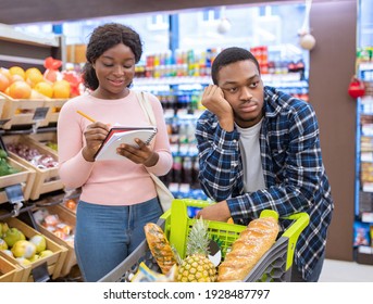 Young black guy feeling bored and tired of shopping for food, his wife checking groceries list at huge mall. Millennial African American family purchasing foodstuff at supermarket - Powered by Shutterstock