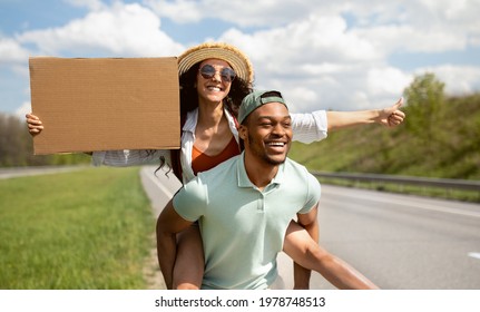 Young black guy giving piggyback ride to his girlfriend, hitchhiking on road together, holding empty sign with mockup outdoors. Happy multiethnic couple traveling by autostop, showing thumb up gesture