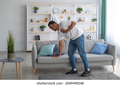 Young Black Guy Having Heart Attack, Holding His Chest In Pain, Leaning On Sofa At Home, Full Length. Millennial African American Man Suffering From Cardiovascular Disease, Getting Infarction Indoors