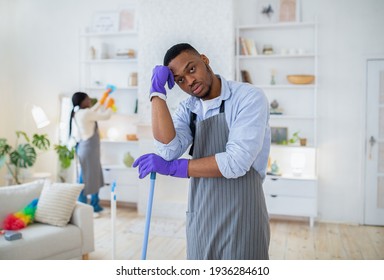 Young black guy leaning on mop, feeling bored or tired of cleaning home with his girlfriend, indoors