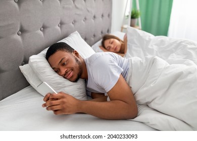 Young Black Guy Lying On Bed And Using Mobile Phone To Text His Lover While His Wife Is Asleep. Millennial Afro Man Having Love Affair, Being Unfaithful To His Girlfriend. Relationship Infidelity