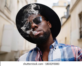 A Young Black Guy In Sunglasses And A Black Hat Blows Cigarette Smoke From His Mouth. A Stylish Smoker In A Plaid Shirt Blows Smoke Directly Into The Camera.