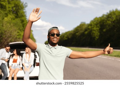 Young black guy trying to stop passing auto, his friends sitting in car trunk of broken vehicle along road, having accident. African American man hitchhiking on highway, needing assistance - Powered by Shutterstock