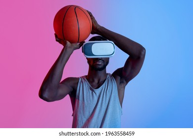 Young Black Guy In VR Headset Throwing Basketball In Neon Light. Motivated Young Sportsman Using Contemporary Technologies For Streetball Workout. Exercising With Virtual Reality Concept