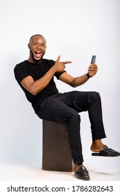 Young Black Handsome Man Sitting, Shocked And Pointing At His Mobile Phone