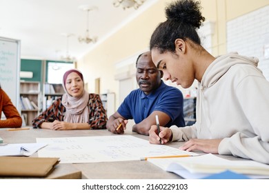 Young Black immigrant sitting at table writing something on handmade educational English language poster during lesson - Shutterstock ID 2160229103