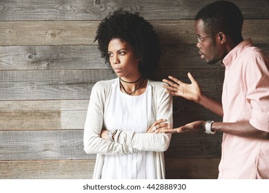 Young black indignant male gesticulating, arguing with his dark skinned girlfriend, who is standing against wooden wall with crossed arms, looking away with deeply offended expression on her face - Powered by Shutterstock