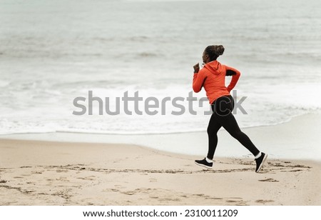 Young black lady athlete in sportswear and headphones running on sea beach in summer, full length. Sports outdoor, training alone, exercise for weight loss, body care and active lifestyle
