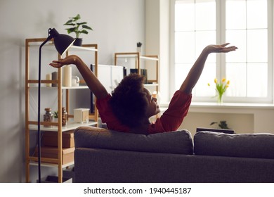 Young black lady enjoying weekend and relaxing on comfortable couch at home. African American woman sitting on sofa in living-room, stretching arms and smiling feeling untroubled and happy, back view