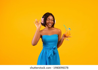 Young Black Lady In Stylish Outfit Holding Exotic Cocktail And Enjoying Favorite Music In Headphones On Orange Background. Posh African American Woman With Tropical Drink Listening To Summer Songs