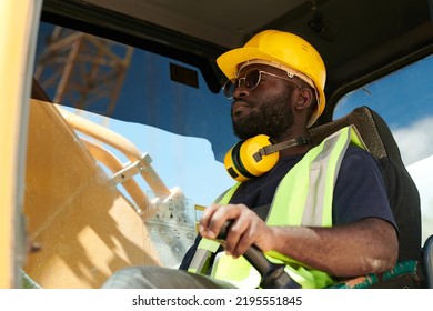 Young Black Male Driver Of Construction Machine In Safety Helmet And Uniform Sitting By Steer And Looking Through Front Window