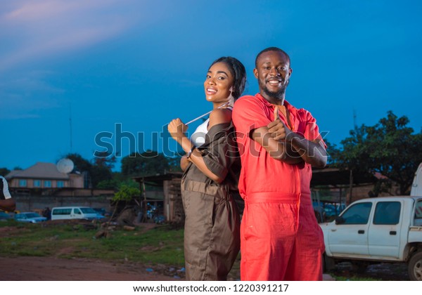 young black male and female engineers smiling and giving\
a thumbs up while standing back to back at a mechanic site workshop\
