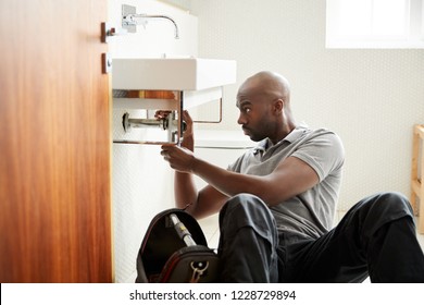 Young Black Male Plumber Sitting On The Floor Fixing A Bathroom Sink, Seen From Doorway