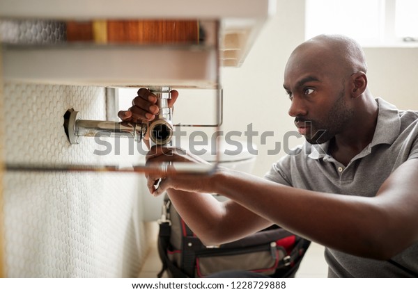 Young black male plumber sitting on the floor\
fixing a bathroom sink, close\
up