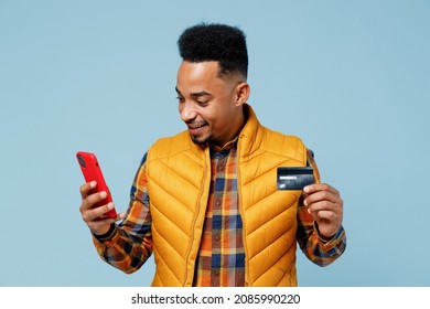 Young Black Man 20s Years Old Wears Yellow Waistcoat Shirt Using Mobile Cell Phone Hold In Hand Credit Bank Card Doing Online Shopping Isolated On Plain Pastel Light Blue Background Studio Portrait