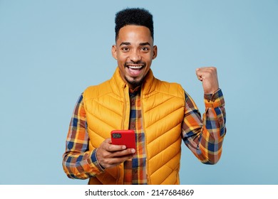 Young black man 20s years old wears yellow waistcoat shirt hold in hand use mobile cell phone doing winner gesture clenching fist say yes isolated on plain pastel light blue background studio portrait