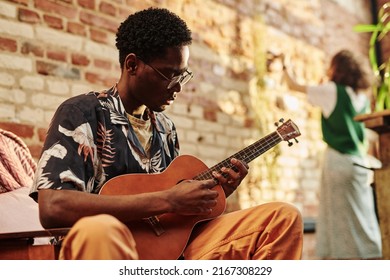 Young Black Man With Acoustic Guitar Sitting In Armchair Against His Girlfriend Standing In Front Of Brick Wall In Living Room Of Loft Apartment