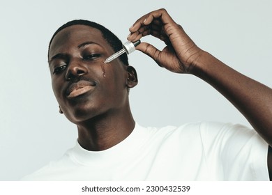 Jeune homme noir appliquant du sérum de beauté sur son visage. Un jeune homme confiant s'occupe de sa peau de mélanine, utilisant un produit nourrissant pour maintenir un teint sain, jeune et sans défaut. : photo de stock